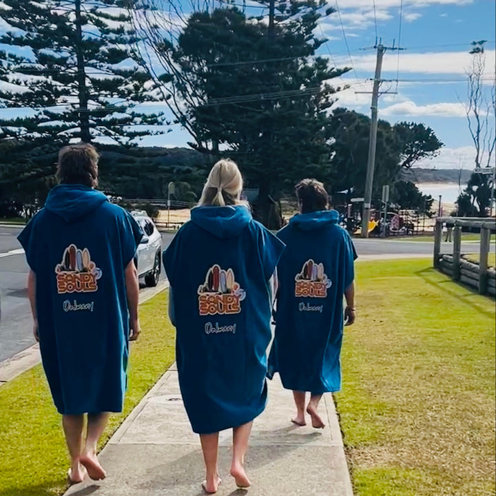 Three people wearing blue robes with a logo and text, walking on a sidewalk near a road with trees and a cloudy sky.