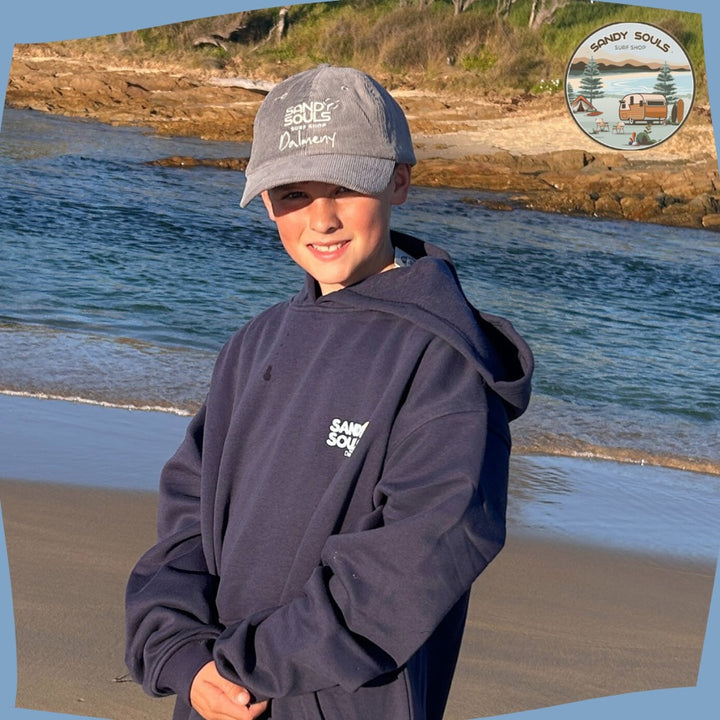 Boy in front of Dalmeny inlet wearing Sandy souls surf shop DALMENy printed navy hooded jumper and grey corduroy cap with Sandy souls surf shop on. 