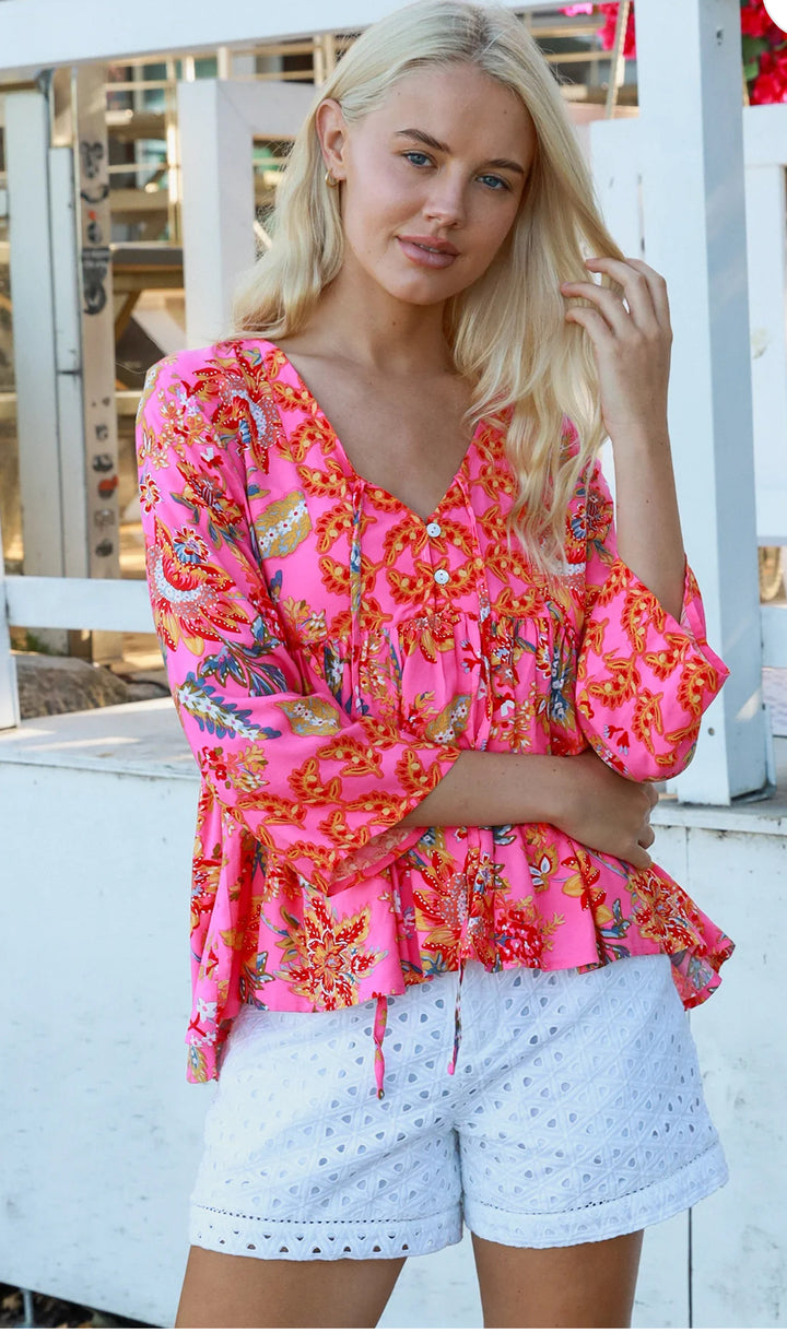 Woman wearing a bright pink floral blouse and white shorts on a wooden deck.