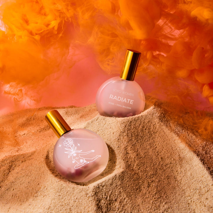 Two pink perfume bottles with gold caps on a sandy surface with orange smoke in the background.