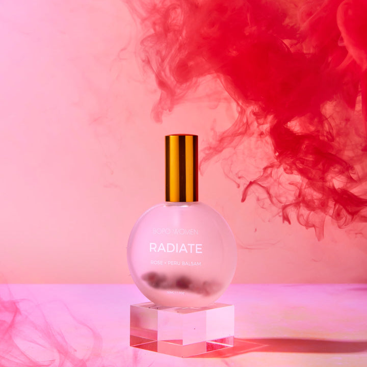 Perfume bottle labeled 'Radiate' with red smoke effect on a pink background