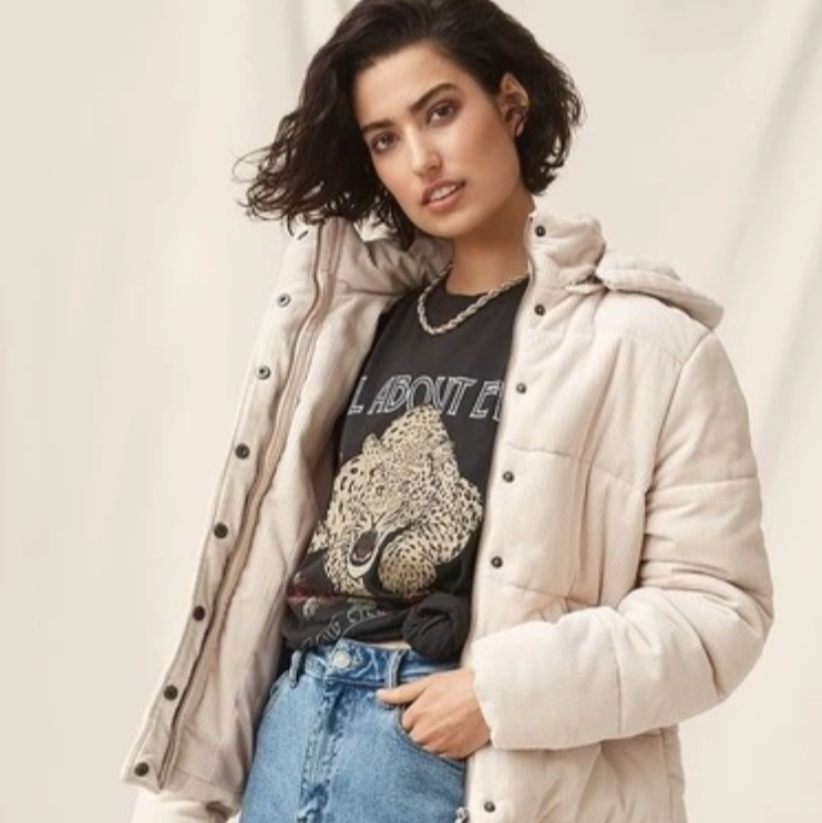 Woman wearing a beige puffer jacket, graphic t-shirt, and blue jeans on a plain background