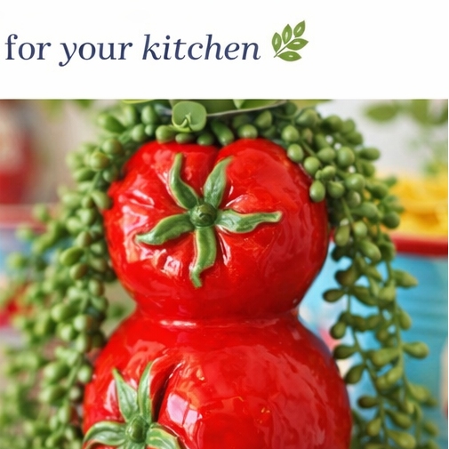 Red ceramic tomato-shaped container with green leaves on a wooden surface, surrounded by plants.